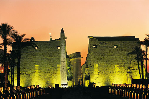 The illuminated entrance of Luxor Temple at night, featuring a towering pink granite obelisk and colossal seated statues of Pharaoh Ramses II under a dark blue sky.